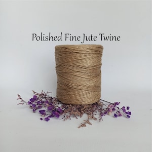 May include: A spool of natural brown jute twine, also known as string or cord, is displayed on a white background. The twine is wrapped around itself and is sitting on top of a bed of purple flowers.