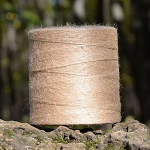 May include: A spool of natural brown twine, made from jute fiber, is sitting on a rough, textured surface. The twine is tightly wound and has a slightly fuzzy texture.