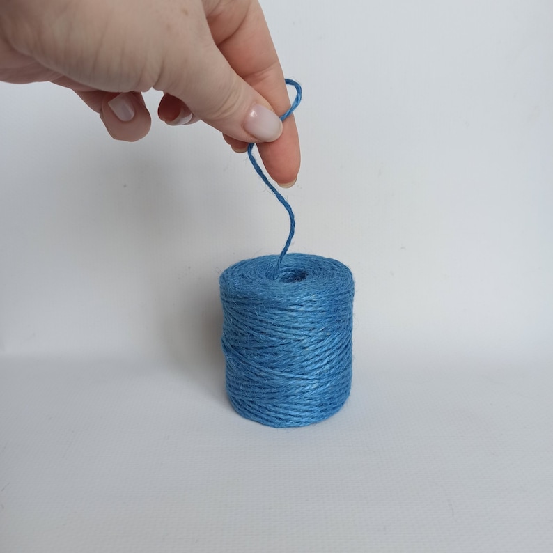 Op de afbeelding: Een spoel blauw touw. Het touw is helderblauw en strak gewikkeld rond een cilindrische kern. Een lus van het touw wordt door een hand omhoog gehouden, wat suggereert dat het wordt gebruikt voor knutselen of verpakken.