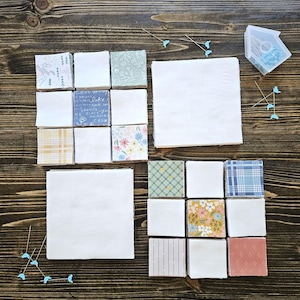 May include: A collection of fabric squares in various patterns and colors, including floral, plaid, and geometric designs. The squares are arranged in a grid pattern on a wooden surface. There are also two larger white fabric squares, one with a pin cushion in the shape of a house.
