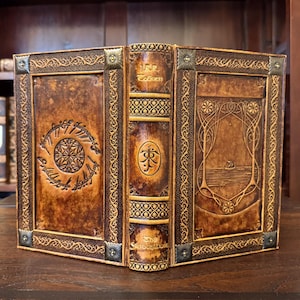 Op de afbeelding: Een handgemaakt, ledergebonden boek met ingewikkelde reli&euml;fontwerpen. De kaft toont een cirkelvormig embleem en schrift, met de naam van de auteur op de rug. Het boek heeft een warme bruine kleur met decoratieve metalen hoekaccenten.