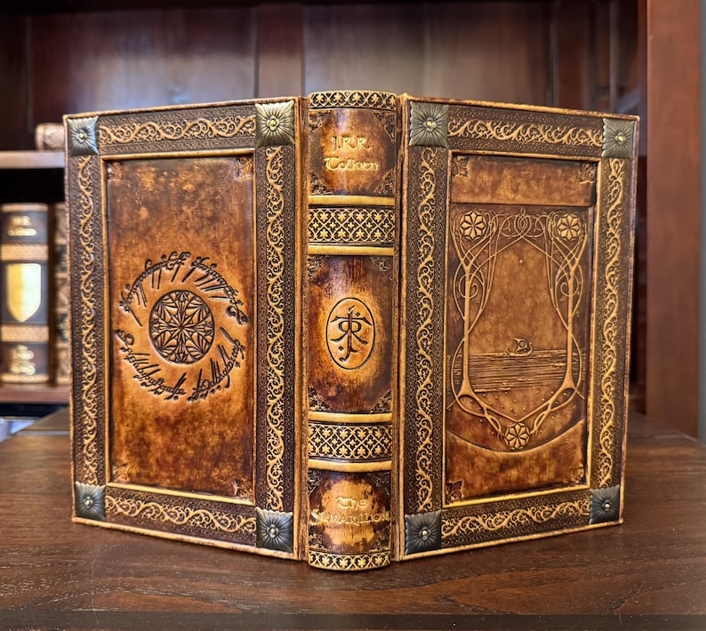 Op de afbeelding: Een handgemaakt, ledergebonden boek met ingewikkelde reli&euml;fontwerpen. De kaft toont een cirkelvormig embleem en schrift, met de naam van de auteur op de rug. Het boek heeft een warme bruine kleur met decoratieve metalen hoekaccenten.