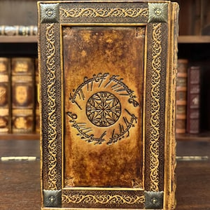 Op de afbeelding: Een ledergebonden boek met een gedetailleerd reli&euml;fontwerp. De kaft is warmbruin met een cirkelvormig embleem en schrift. Sierlijke goudkleurige randen en hoekaccenten omlijsten de kaft. Het boek staat op een houten oppervlak.