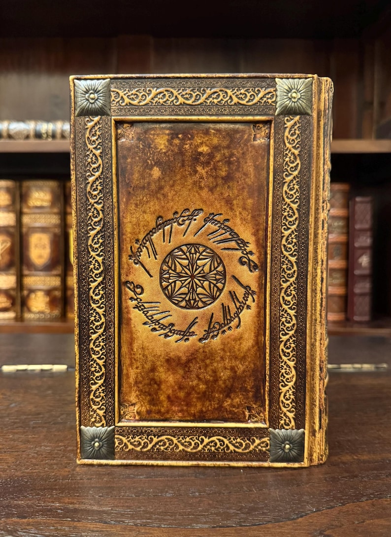 Op de afbeelding: Een ledergebonden boek met een gedetailleerd reli&euml;fontwerp. De kaft is warmbruin met een cirkelvormig embleem en schrift. Sierlijke goudkleurige randen en hoekaccenten omlijsten de kaft. Het boek staat op een houten oppervlak.