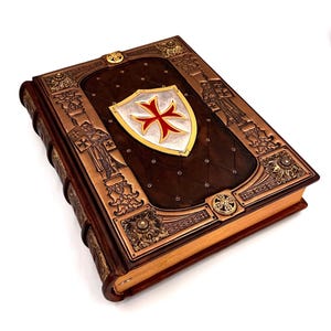 May include: A brown leather-bound book with a decorative copper-coloured frame and a shield emblem. The shield features a red cross on a white background, with a gold border. The book's cover has a diamond-quilted pattern.