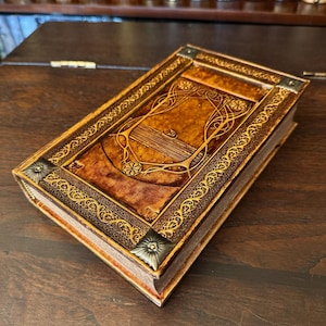 Op de afbeelding: Een handgemaakt, ledergebonden notitieboek met een reli&euml;f omslag met een Art Nouveau-ontwerp. Het notitieboek heeft een rijke, bruine kleur met gouden accenten en decoratieve hoekstukken. De pagina's zijn zichtbaar, wat duidt op een nieuw of weinig gebruikt boek.