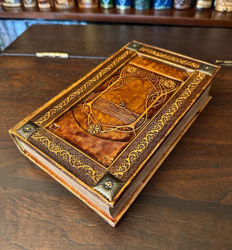 Op de afbeelding: Een handgemaakt, ledergebonden notitieboek met een reli&euml;f omslag met een Art Nouveau-ontwerp. Het notitieboek heeft een rijke, bruine kleur met gouden accenten en decoratieve hoekstukken. De pagina's zijn zichtbaar, wat duidt op een nieuw of weinig gebruikt boek.
