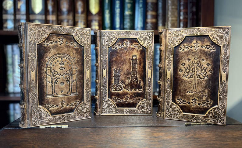 K&ouml;nnte beinhalten: Drei ledergebundene B&uuml;cher mit aufwendigen Designs. Die Einb&auml;nde zeigen gepr&auml;gte Bilder einer T&uuml;r, eines Turms und eines Baumes, alle in Gold- und Braunt&ouml;nen. Die B&uuml;cher stehen auf einer Holzoberfl&auml;che, mit einem verschwommenen B&uuml;cherregal im Hintergrund.