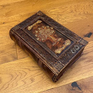 May include: A brown, rectangular box with ornate detailing and a gold-colored tree design on the top. The box has a leather-like appearance with embossed patterns and a decorative border. The box is resting on a wooden surface.