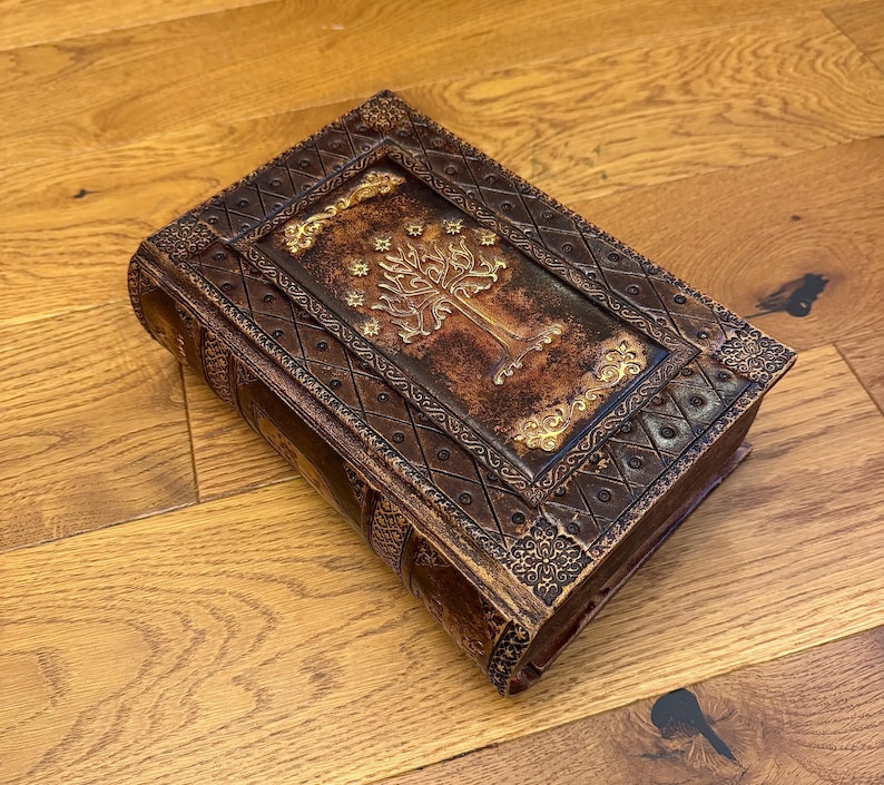May include: A brown, rectangular box with ornate detailing and a gold-colored tree design on the top. The box has a leather-like appearance with embossed patterns and a decorative border. The box is resting on a wooden surface.
