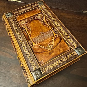 Op de afbeelding: Een gedetailleerd boek in antieke stijl met een bruine leren kaft. De kaft heeft reli&euml;f bloemen- en geometrische ontwerpen, met gouden accenten en metalen hoekstukken. De randen van het boek zijn ook goudkleurig.