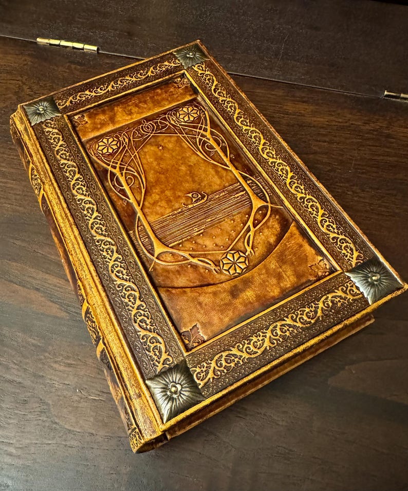 Op de afbeelding: Een gedetailleerd boek in antieke stijl met een bruine leren kaft. De kaft heeft reli&euml;f bloemen- en geometrische ontwerpen, met gouden accenten en metalen hoekstukken. De randen van het boek zijn ook goudkleurig.