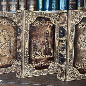 K&ouml;nnte beinhalten: Drei ledergebundene B&uuml;cher mit aufwendigen Designs. Die Einb&auml;nde zeigen gepr&auml;gte Bilder eines Tores, eines Turms und eines Baumes, in Braun- und Goldt&ouml;nen. Die B&uuml;cher stehen auf einem Holzregal, was auf ein Fantasy- oder historisches Thema hindeutet.