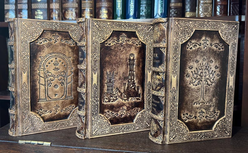 K&ouml;nnte beinhalten: Drei ledergebundene B&uuml;cher mit aufwendigen Designs. Die Einb&auml;nde zeigen gepr&auml;gte Bilder eines Tores, eines Turms und eines Baumes, in Braun- und Goldt&ouml;nen. Die B&uuml;cher stehen auf einem Holzregal, was auf ein Fantasy- oder historisches Thema hindeutet.