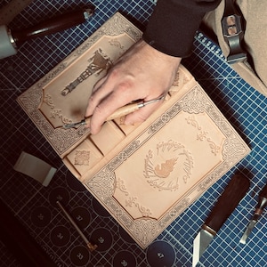 K&ouml;nnte beinhalten: Ein hellbraunes Lederjournal mit aufwendigen gepr&auml;gten Designs und einem zentralen Emblem. Das Journal ist ge&ouml;ffnet, eine Hand verwendet ein Schnitzwerkzeug, um Details hinzuzuf&uuml;gen. Werkzeuge und eine Schneidematte sind im Hintergrund.