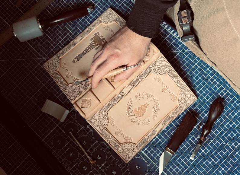 K&ouml;nnte beinhalten: Ein hellbraunes Lederjournal mit aufwendigen gepr&auml;gten Designs und einem zentralen Emblem. Das Journal ist ge&ouml;ffnet, eine Hand verwendet ein Schnitzwerkzeug, um Details hinzuzuf&uuml;gen. Werkzeuge und eine Schneidematte sind im Hintergrund.