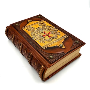 Könnte beinhalten: Ein braunes, ledergebundenes Tagebuch mit aufwendigen goldenen Verzierungen und einem roten Kreuz auf dem Einband. Das Tagebuch hat ein mittelalterliches Design und ist perfekt zum Schreiben oder zum Aufbewahren von Geheimnissen.