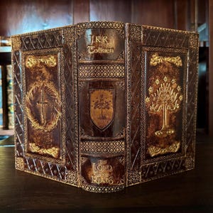 May include: A decorative book featuring the title "The Lord of the Rings" by J.R.R. Tolkien. The book has a brown leather cover with gold embossed designs, including a cross, a tree, and a shield. The book is displayed on a dark wooden surface.