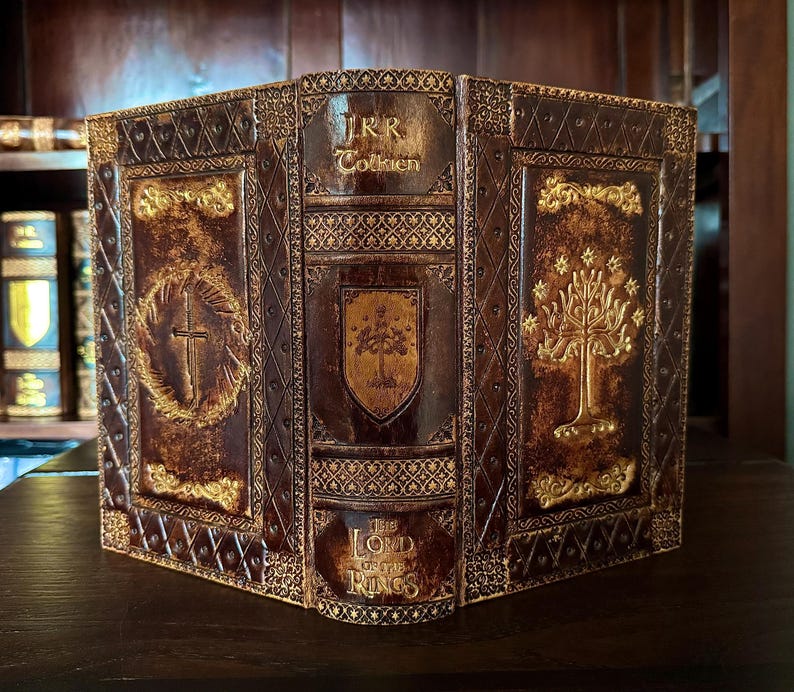 May include: A decorative book featuring the title "The Lord of the Rings" by J.R.R. Tolkien. The book has a brown leather cover with gold embossed designs, including a cross, a tree, and a shield. The book is displayed on a dark wooden surface.