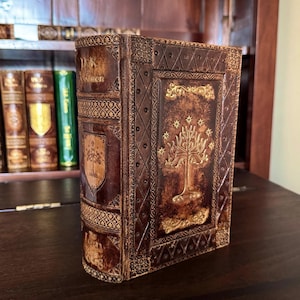 May include: A brown leather-bound book with gold embossed details. The cover features a tree design and the title "The Lord of the Rings". The book has decorative patterns and a shield emblem. The book is displayed on a dark wooden surface.