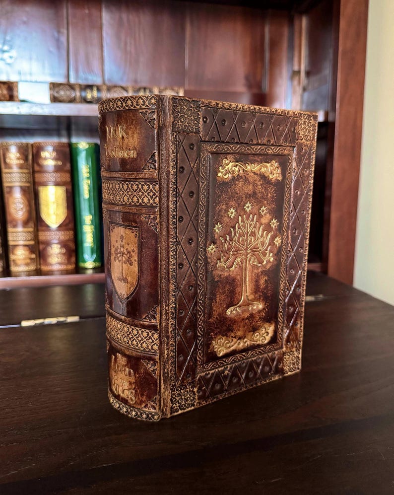 May include: A brown leather-bound book with gold embossed details. The cover features a tree design and the title "The Lord of the Rings". The book has decorative patterns and a shield emblem. The book is displayed on a dark wooden surface.