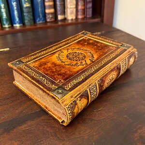 Op de afbeelding: Een decoratief boek met een bruine en gouden kaft met een ingewikkeld ontwerp en tekst. Het boek ligt op een donker houten oppervlak, met een boekenplank op de achtergrond.
