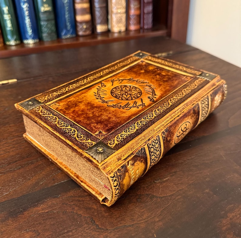 Op de afbeelding: Een decoratief boek met een bruine en gouden kaft met een ingewikkeld ontwerp en tekst. Het boek ligt op een donker houten oppervlak, met een boekenplank op de achtergrond.