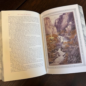 K&ouml;nnte beinhalten: Ein offenes Buch mit einer ganzseitigen Illustration einer Berglandschaft mit einem Wasserfall. Die Seiten des Buches sind cremefarben und der Text ist schwarz. Die Illustration ist mit einem dekorativen Rahmen versehen. Das Buch liegt auf einer dunklen Holzoberfl&auml;che.