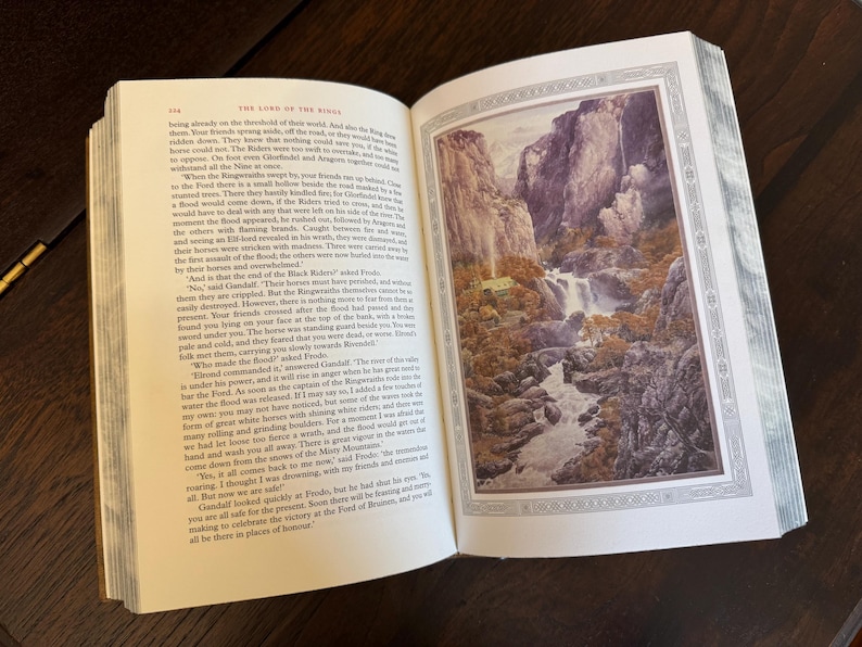 K&ouml;nnte beinhalten: Ein offenes Buch mit einer ganzseitigen Illustration einer Berglandschaft mit einem Wasserfall. Die Seiten des Buches sind cremefarben und der Text ist schwarz. Die Illustration ist mit einem dekorativen Rahmen versehen. Das Buch liegt auf einer dunklen Holzoberfl&auml;che.