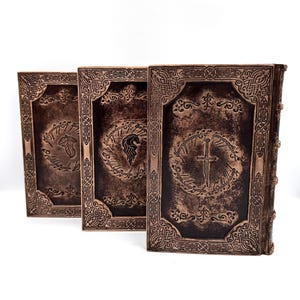 K&ouml;nnte beinhalten: Drei ledergebundene B&uuml;cher mit gepr&auml;gten Designs. Die Einb&auml;nde sind dunkelbraun mit goldenen Akzenten und weisen komplizierte Muster und Symbole auf. Die B&uuml;cher sind nebeneinander angeordnet, wobei die Buchr&uuml;cken zum Betrachter zeigen.