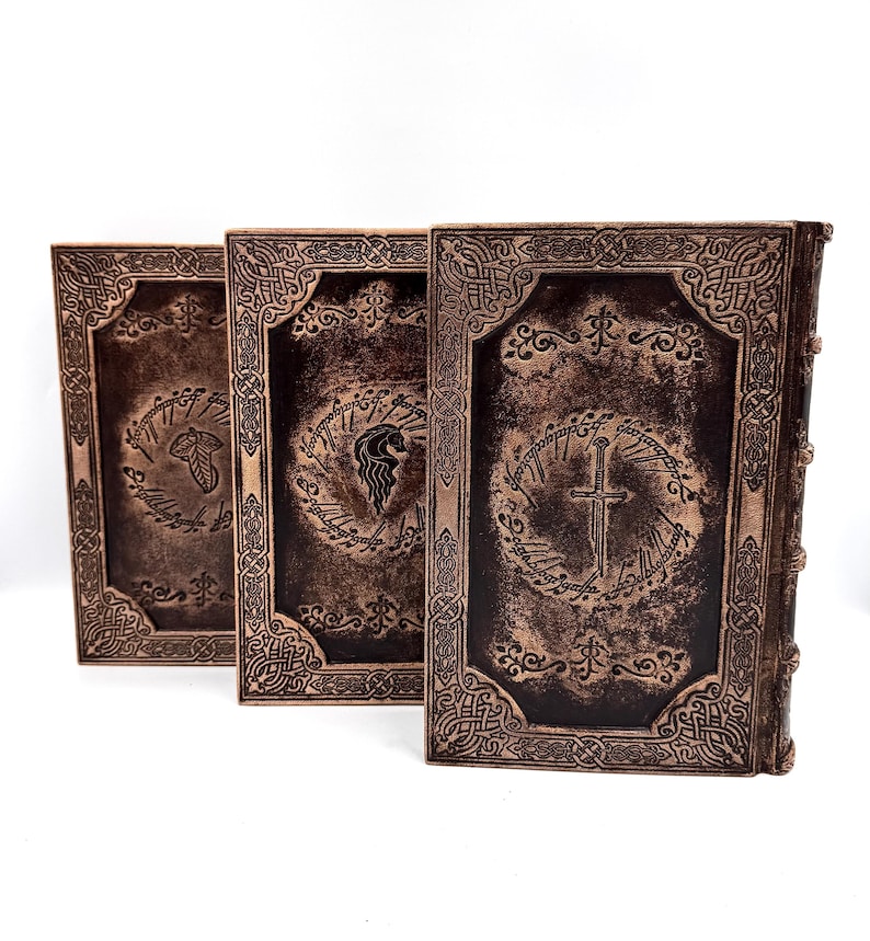 K&ouml;nnte beinhalten: Drei ledergebundene B&uuml;cher mit gepr&auml;gten Designs. Die Einb&auml;nde sind dunkelbraun mit goldenen Akzenten und weisen komplizierte Muster und Symbole auf. Die B&uuml;cher sind nebeneinander angeordnet, wobei die Buchr&uuml;cken zum Betrachter zeigen.