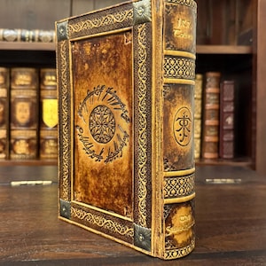 Op de afbeelding: Een bruin leren boek met gouden accenten en ingewikkelde ontwerpen. De kaft heeft een cirkelvormig embleem en tekst, met decoratieve hoekstukken. De rug heeft tekst en een symbool. Het boek staat op een houten oppervlak.