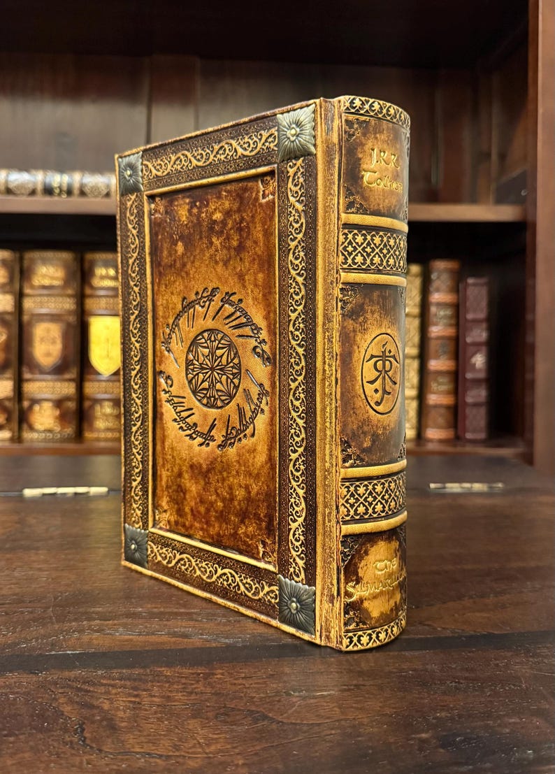 Op de afbeelding: Een bruin leren boek met gouden accenten en ingewikkelde ontwerpen. De kaft heeft een cirkelvormig embleem en tekst, met decoratieve hoekstukken. De rug heeft tekst en een symbool. Het boek staat op een houten oppervlak.