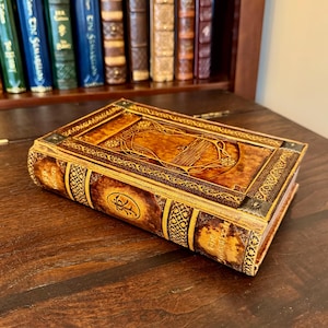 Op de afbeelding: Een decoratieve, boekvormige doos met een gedetailleerd, reli&euml;f ontwerp. De doos is voornamelijk bruin met gouden accenten en sierlijke patronen. Het rust op een donker houten oppervlak, met een boekenkast op de achtergrond.