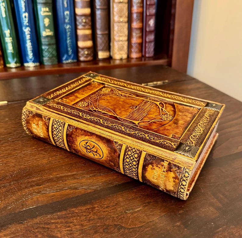 Op de afbeelding: Een decoratieve, boekvormige doos met een gedetailleerd, reli&euml;f ontwerp. De doos is voornamelijk bruin met gouden accenten en sierlijke patronen. Het rust op een donker houten oppervlak, met een boekenkast op de achtergrond.