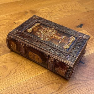 May include: A brown, leather-bound book with intricate gold embossed detailing. The cover features a tree design with ornate borders. The book's edges and spine are also decorated with embossed patterns, creating a vintage aesthetic.