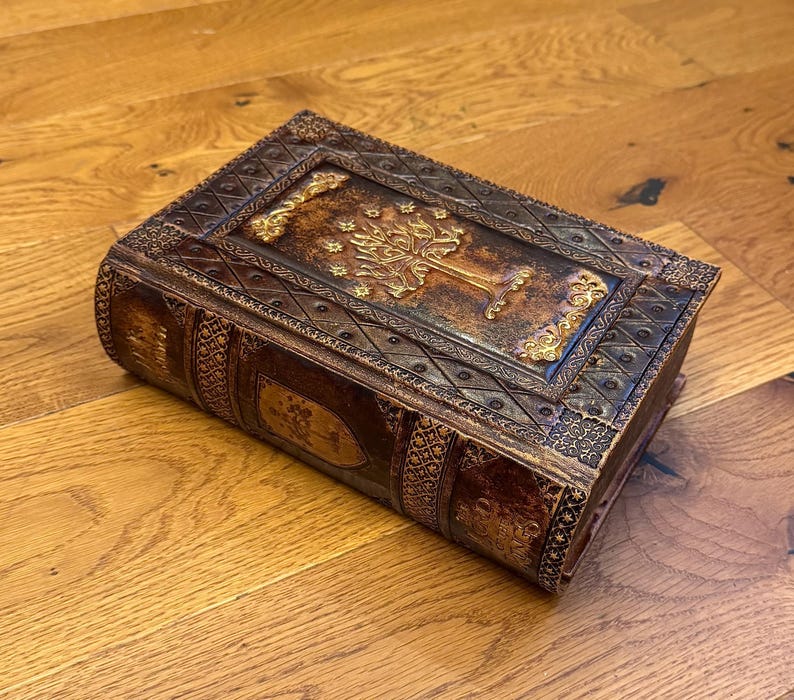 May include: A brown, leather-bound book with intricate gold embossed detailing. The cover features a tree design with ornate borders. The book's edges and spine are also decorated with embossed patterns, creating a vintage aesthetic.