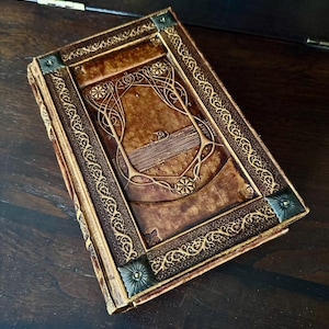 Op de afbeelding: Een bruin leren boek met ingewikkelde goudkleurige reli&euml;fdesigns. De kaft heeft een centraal rechthoekig paneel met bloemen- en geometrische patronen, omlijst door een decoratieve rand. De hoeken zijn versierd met metalen accenten.