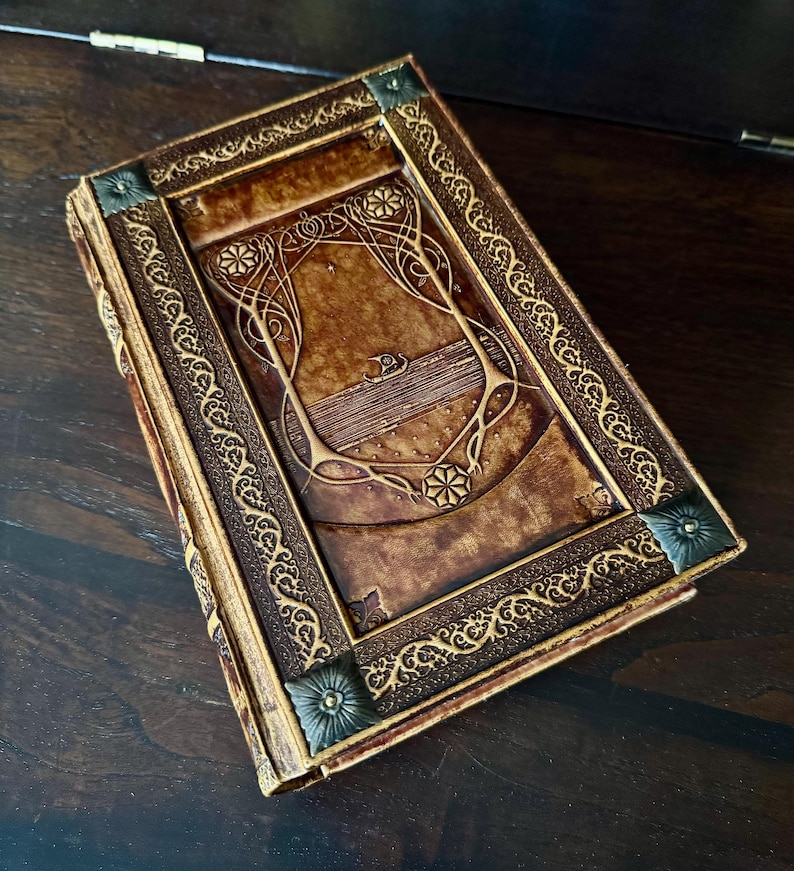 Op de afbeelding: Een bruin leren boek met ingewikkelde goudkleurige reli&euml;fdesigns. De kaft heeft een centraal rechthoekig paneel met bloemen- en geometrische patronen, omlijst door een decoratieve rand. De hoeken zijn versierd met metalen accenten.