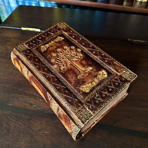 May include: A brown leather-bound journal with a gold embossed tree design on the cover. The journal has decorative gold accents and a diamond pattern. The pages are visible, suggesting it is a new, unused book.