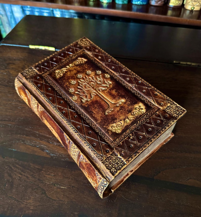 May include: A brown leather-bound journal with a gold embossed tree design on the cover. The journal has decorative gold accents and a diamond pattern. The pages are visible, suggesting it is a new, unused book.