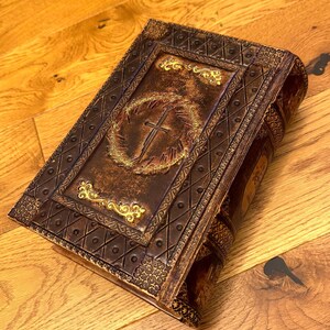 May include: A decorative, antique-style book with a brown leather cover. The cover features gold-colored filigree and a central design of a cross within a wreath. The book has a detailed, embossed design and a worn, vintage appearance.