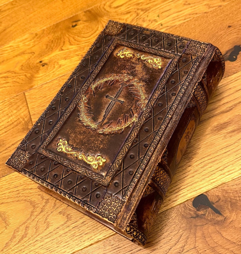 May include: A decorative, antique-style book with a brown leather cover. The cover features gold-colored filigree and a central design of a cross within a wreath. The book has a detailed, embossed design and a worn, vintage appearance.
