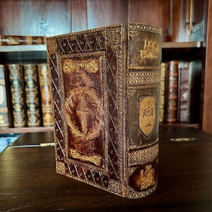 May include: A decorative book with a brown and gold cover. The cover features a cross within a wreath and the text "The Lord of the Rings". The book has an aged, antique appearance, with intricate embossed designs and a shield emblem.