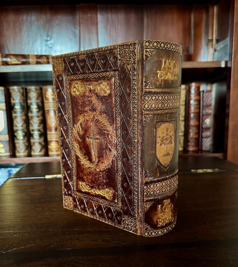 May include: A decorative book with a brown and gold cover. The cover features a cross within a wreath and the text "The Lord of the Rings". The book has an aged, antique appearance, with intricate embossed designs and a shield emblem.