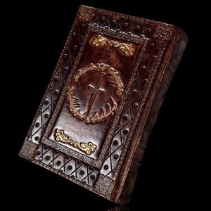 May include: A brown leather-bound book with ornate gold detailing. The cover features a cross within a wreath and decorative patterns. The book's edges and corners are also embellished with gold accents, creating a vintage aesthetic.