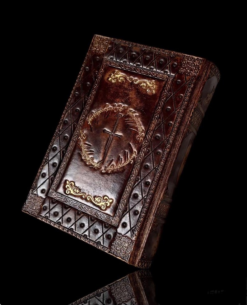 May include: A brown leather-bound book with ornate gold detailing. The cover features a cross within a wreath and decorative patterns. The book's edges and corners are also embellished with gold accents, creating a vintage aesthetic.