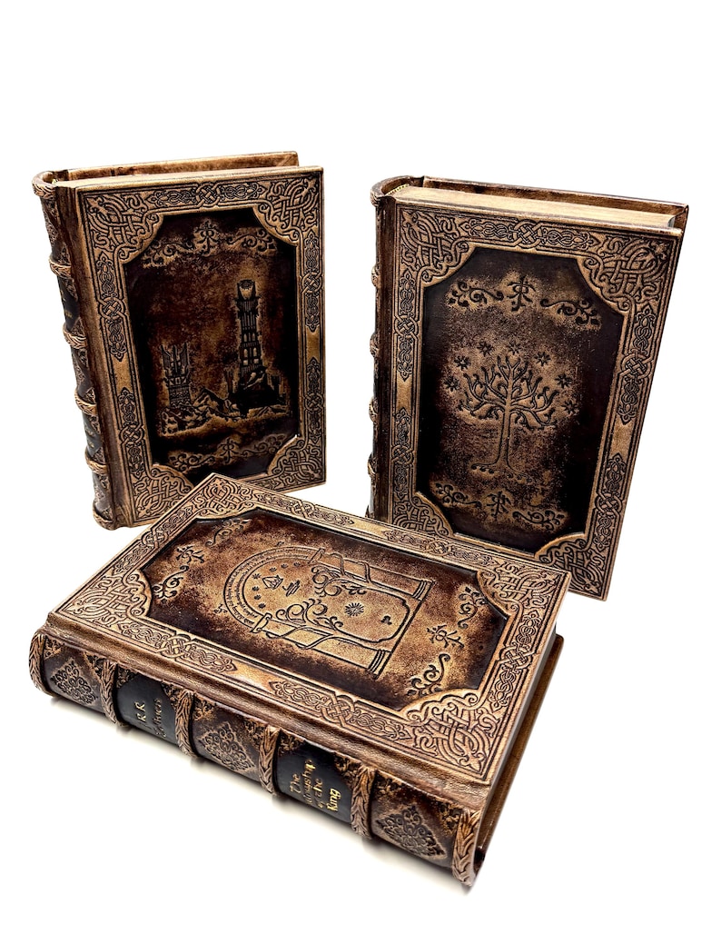K&ouml;nnte beinhalten: Drei braune dekorative B&uuml;cher mit aufwendigen gepr&auml;gten Designs. Die Einb&auml;nde zeigen Szenen eines Turms, eines Baumes und einer Karte. Die Buchr&uuml;cken haben dekorative B&auml;nder und den Text "The Fellowship of the Ring" und "The Return of the King".