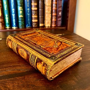 Op de afbeelding: Een decoratief boek in antieke stijl met een bruine leren kaft en gouden accenten. De kaft heeft sierlijke reli&euml;fontwerpen en een rechthoekig middenpaneel. Het boek ligt op een donker houten oppervlak, met een boekenkast op de achtergrond.