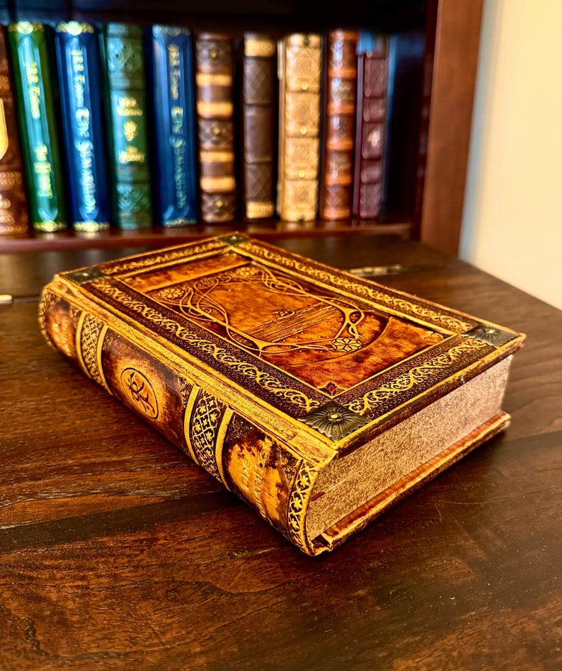 Op de afbeelding: Een decoratief boek in antieke stijl met een bruine leren kaft en gouden accenten. De kaft heeft sierlijke reli&euml;fontwerpen en een rechthoekig middenpaneel. Het boek ligt op een donker houten oppervlak, met een boekenkast op de achtergrond.